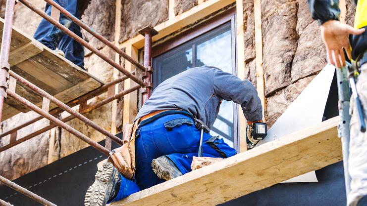 Bauarbeiter installiert Material an einer Hauswand auf einem Gerüst, während ein Kollege am Fenster assistiert.